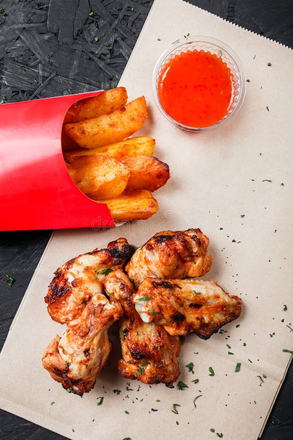 Le ali di pollo fritto con le patate al forno ed il bbq sauce sulla carta del mestiere su fondo nero fotografie stock libere da diritti Le ali di pollo fritto con le patate al forno ed il bbq sauce sulla carta del mestiere su fondo nero fotografie stock libere da diritti