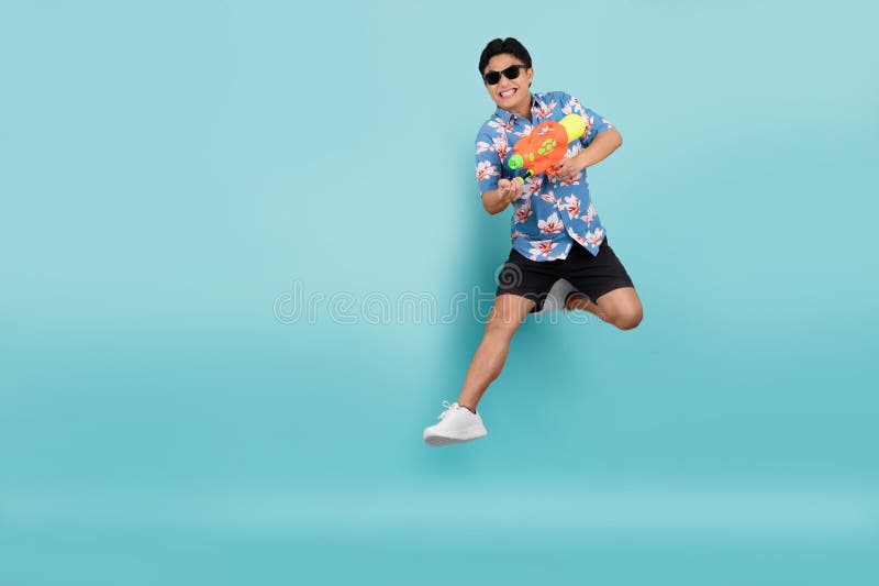 Le beau jeune asiatique en vêtements d'été saute et utilise un pistolet à eau sur fond bleu. festival songkran en thaïlande image stock
