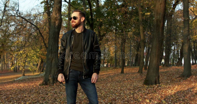 Le beau macho barbu porte des lunettes de soleil regarde autour de lui et sourit dans le parc
