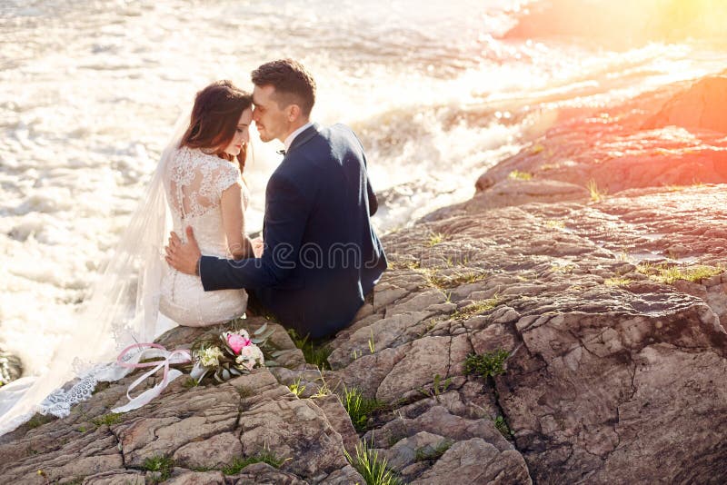 Le belle coppie amano baciare mentre si siedono sulle rocce si avvicinano al fiume immagine stock Le belle coppie amano baciare mentre si siedono sulle rocce si avvicinano al fiume immagine stock