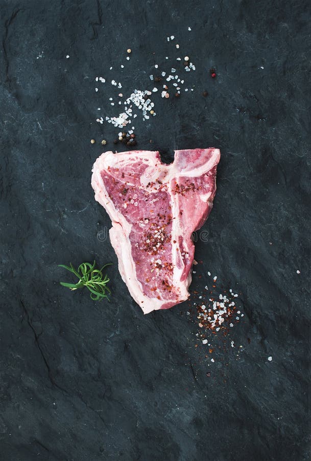 Le bifteck de boeuf frais cru d'à l'os avec du sel, les épices et le romarin au-dessus de l'ardoise noire lapident le fond, copie photographie stock libre de droits Le bifteck de boeuf frais cru d'à l'os avec du sel, les épices et le romarin au-dessus de l'ardoise noire lapident le fond, copie photographie stock libre de droits