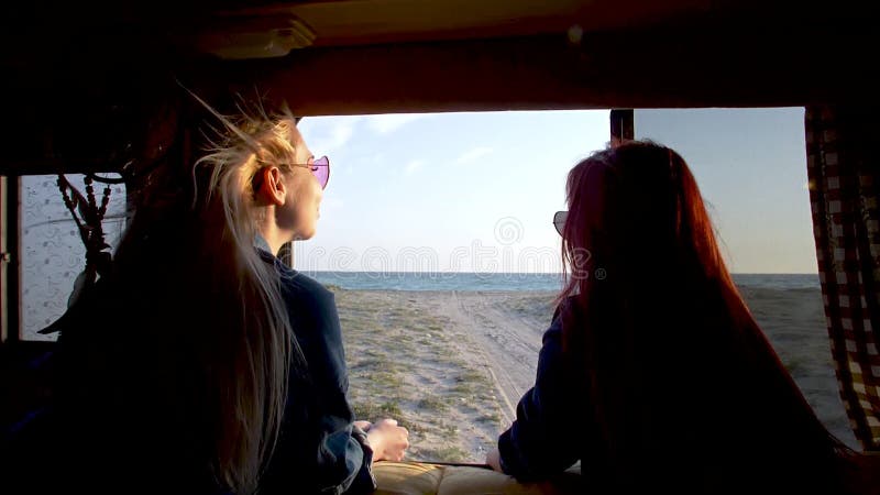Le coppie delle giovani donne esaminano alla spiaggia sabbiosa ed al mare attraverso la finestra automatica del rimorchio i capel