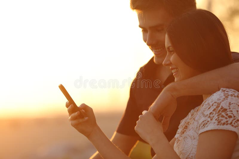 Le coppie facendo uso di uno smartphone in un tramonto indietro si accendono immagini stock libere da diritti Le coppie facendo uso di uno smartphone in un tramonto indietro si accendono immagini stock libere da diritti
