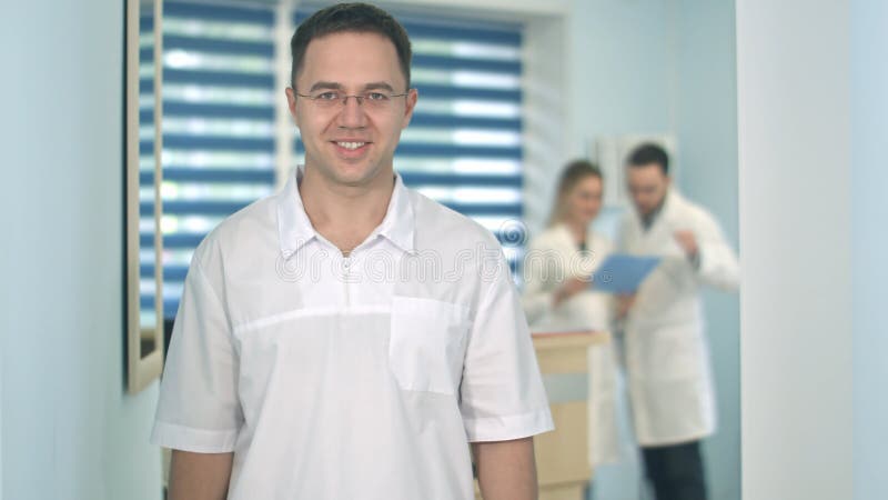 Le den manliga doktorn i exponeringsglas som ser kameran medan medicinsk personal som arbetar på bakgrunden arkivbild Le den manliga doktorn i exponeringsglas som ser kameran medan medicinsk personal som arbetar på bakgrunden arkivbild