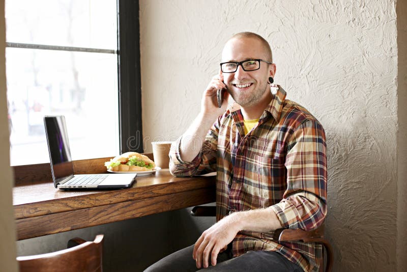 Le den skäggiga affärsmannen som bär tillfälliga hipsterkläder genom att använda bärbar dator- och cellsmartphonen i coffehus arkivbilder Le den skäggiga affärsmannen som bär tillfälliga hipsterkläder genom att använda bärbar dator- och cellsmartphonen i coffehus arkivbilder