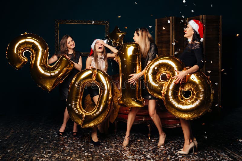 Group of cheerful young women in Christmas hats holding gold colored numbers and throwing confetti at New Year party. Group of cheerful young women in Christmas hats holding gold colored numbers and throwing confetti at New Year party