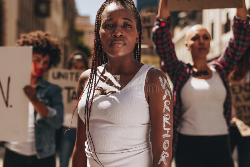 Young women standing outdoors with group of protesters at back. Woman with word warrior written on her arm. Young women standing outdoors with group of protesters at back. Woman with word warrior written on her arm.