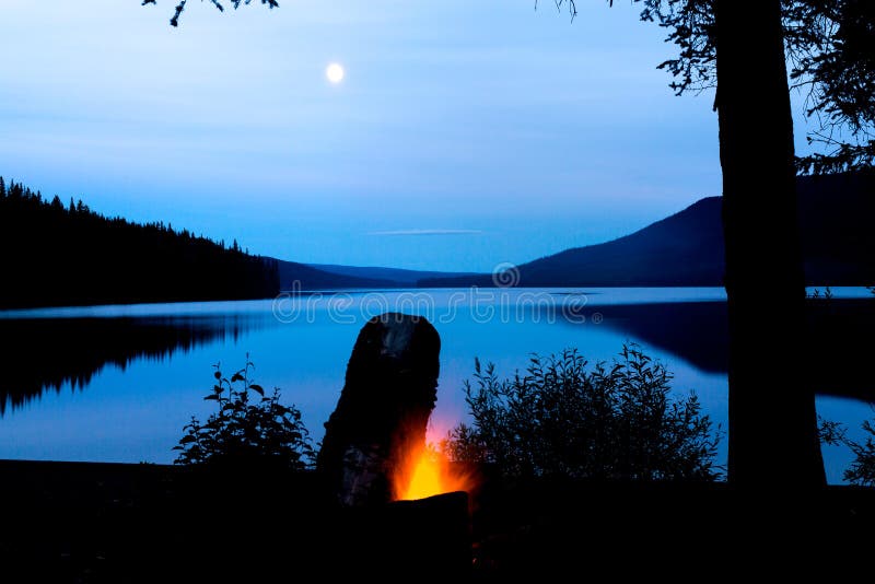 A full moon rises over a still lake during the night. In the foreground a campfire rages with a fresh stock of dry firewood. A full moon rises over a still lake during the night. In the foreground a campfire rages with a fresh stock of dry firewood.