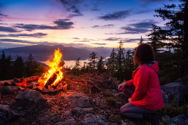 Adventure Girl by the Campfire on top of a mountain with Beautiful Canadian Nature Landscape in background during a colorful Sunset. Taken on Bowen Island, near Vancouver, British Columbia, Canada. Adventure Girl by the Campfire on top of a mountain with Beautiful Canadian Nature Landscape in background during a colorful Sunset. Taken on Bowen Island, near Vancouver, British Columbia, Canada.