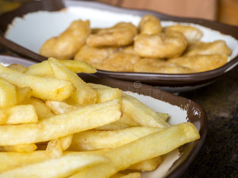 Le fritture di Fried French e le pepite di pollo su un piatto bianco con marrone rasentano la tavola di legno fotografia stock libera da diritti Le fritture di Fried French e le pepite di pollo su un piatto bianco con marrone rasentano la tavola di legno fotografia stock libera da diritti