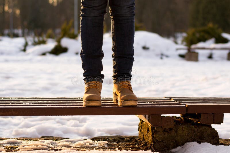 Le gambe femminili stanno stando sul banco nel giorno soleggiato dell'inverno Fondo vago dei raggi di tramonto e della neve Girk immagini stock Le gambe femminili stanno stando sul banco nel giorno soleggiato dell'inverno Fondo vago dei raggi di tramonto e della neve Girk immagini stock