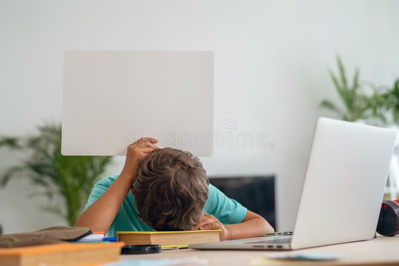 Le garçon assez triste est assis à la table avec le sort des livres et tient le signe blanc photos libres de droits