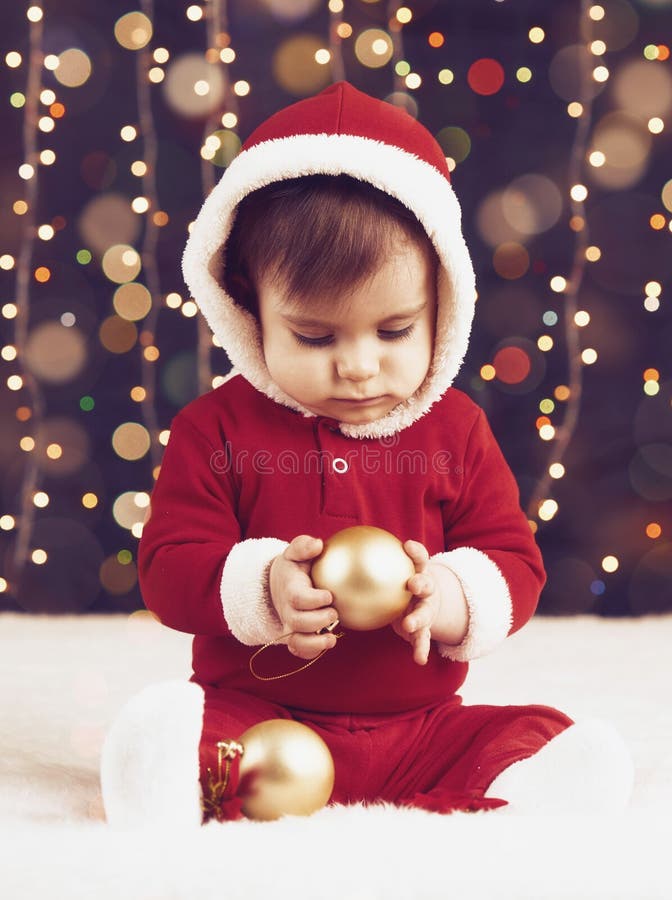 Le garçon de petit enfant habillé comme Santa jouant avec la décoration de Noël, le fond foncé avec l'illumination et le boke s'a photographie stock Le garçon de petit enfant habillé comme Santa jouant avec la décoration de Noël, le fond foncé avec l'illumination et le boke s'a photographie stock