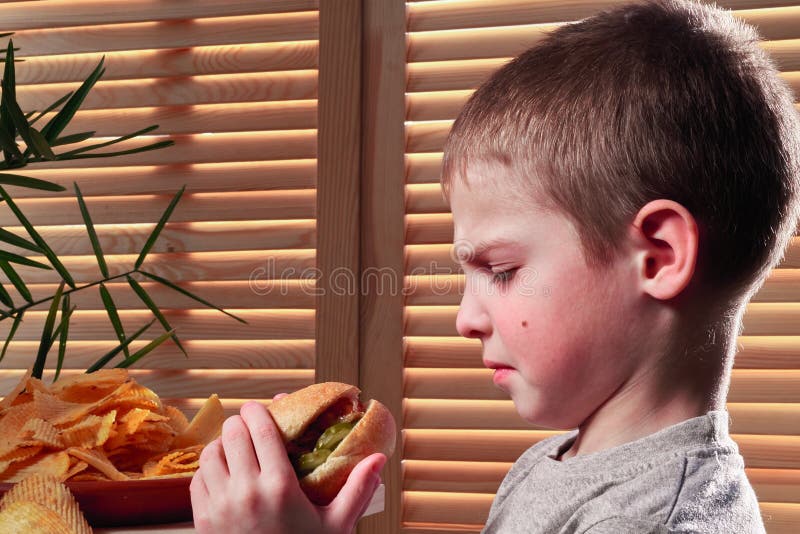 Le garçon fronçant les sourcils regarde le hot dog L'enfant regarde fixement avec dégoût la nourriture insipide dans le café Alim images libres de droits Le garçon fronçant les sourcils regarde le hot dog L'enfant regarde fixement avec dégoût la nourriture insipide dans le café Alim images libres de droits