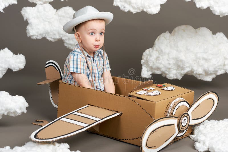 Le garçon joue dans un avion fait de boîte en carton et rêves de devenir un pilote, nuages de l'ouate sur un fond gris, r photo libre de droits Le garçon joue dans un avion fait de boîte en carton et rêves de devenir un pilote, nuages de l'ouate sur un fond gris, r photo libre de droits