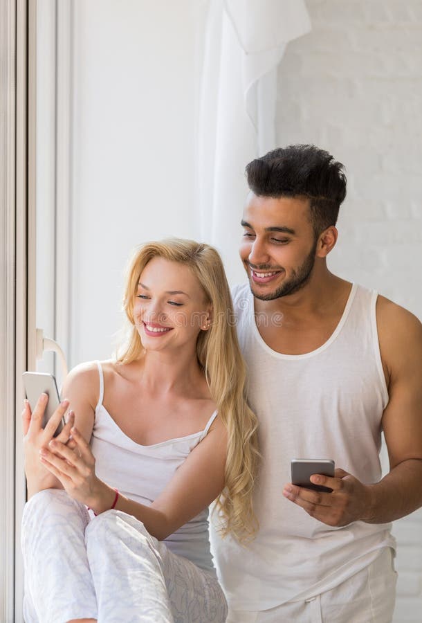 Le giovani belle coppie stanno vicino alla grande finestra, facendo uso della donna ispana dell'uomo di sorriso felice dello Smar fotografie stock Le giovani belle coppie stanno vicino alla grande finestra, facendo uso della donna ispana dell'uomo di sorriso felice dello Smar fotografie stock