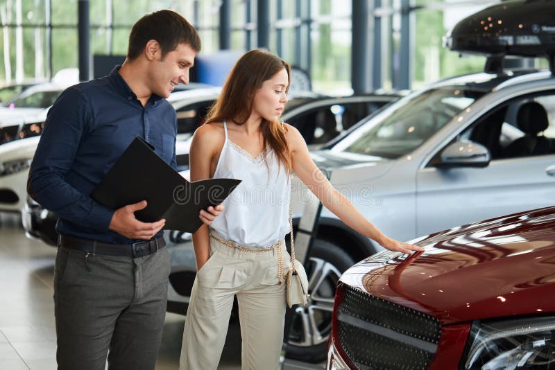 Le giovani coppie in vestiti eleganti di ora legale scelgono una nuova automobile al concessionario auto immagine stock Le giovani coppie in vestiti eleganti di ora legale scelgono una nuova automobile al concessionario auto immagine stock