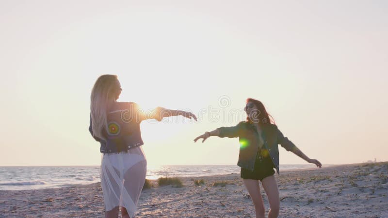 Le giovani donne del movimento lento vestite nello stile di boho stanno divertendo sulla spiaggia nella sera nei raggi del tramon