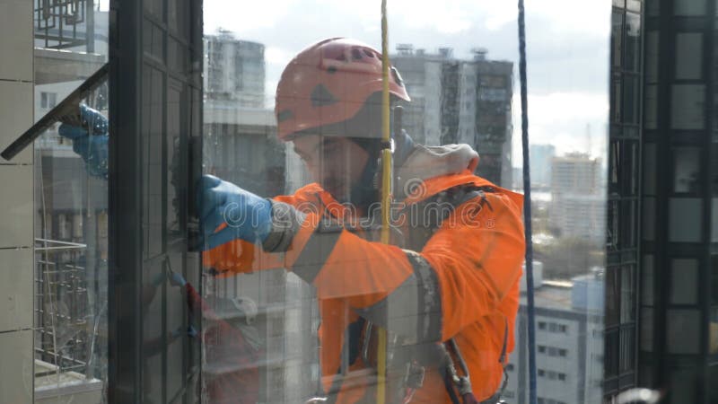 Professional industrial climber in helmet and gloves washes windows with screed view through glass against the background of tall megalopolis buildings, close-up slow motion. Professional industrial climber in helmet and gloves washes windows with screed view through glass against the background of tall megalopolis buildings, close-up slow motion