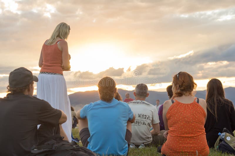 Le groupe de personnes s'asseyant sur l'herbe et apprécient le coucher du soleil photographie stock Le groupe de personnes s'asseyant sur l'herbe et apprécient le coucher du soleil photographie stock