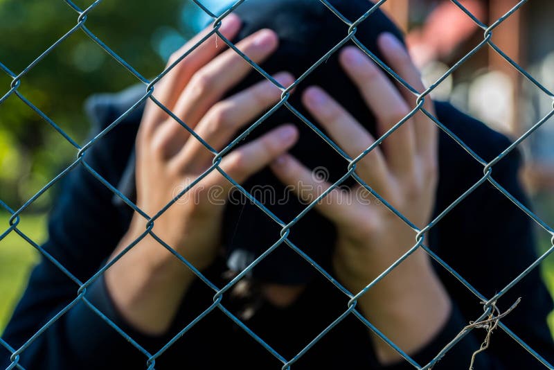 Young unidentifiable teenage boy holding hes head at the correctional institute ,conceptual image of juvenile delinquency, focus on the wired fence. Young unidentifiable teenage boy holding hes head at the correctional institute ,conceptual image of juvenile delinquency, focus on the wired fence.