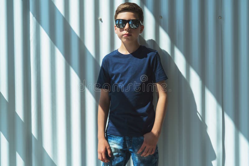 Le jeune garçon de hippie dans des lunettes de soleil habillées dans le T-shirt noir est des positions d'intérieur contre le mur image stock Le jeune garçon de hippie dans des lunettes de soleil habillées dans le T-shirt noir est des positions d'intérieur contre le mur image stock
