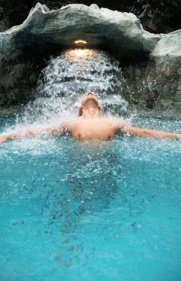 Le jeune homme apprécie l'eau en baisse sous la cascade dans la piscine photographie stock libre de droits Le jeune homme apprécie l'eau en baisse sous la cascade dans la piscine photographie stock libre de droits