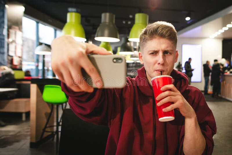 Le jeune homme boit une boisson d'un verre rouge et fait un selfie photo libre de droits Le jeune homme boit une boisson d'un verre rouge et fait un selfie photo libre de droits