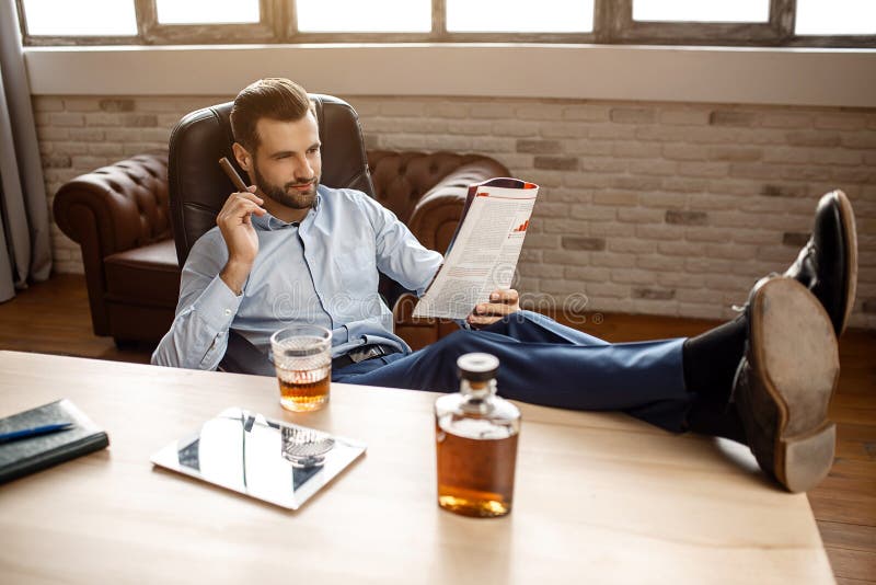 Le jeune homme d'affaires bel s'asseyent sur la chaise et le journal lu dans son propre bureau Il tiennent le cigare à dispositio photo stock Le jeune homme d'affaires bel s'asseyent sur la chaise et le journal lu dans son propre bureau Il tiennent le cigare à dispositio photo stock