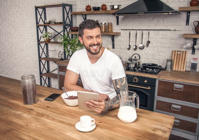 Le jeune homme de sourire bel prend ses céréales de petit déjeuner avec du lait à la cuisine et lit des nouvelles de matin sur le photos libres de droits Le jeune homme de sourire bel prend ses céréales de petit déjeuner avec du lait à la cuisine et lit des nouvelles de matin sur le photos libres de droits