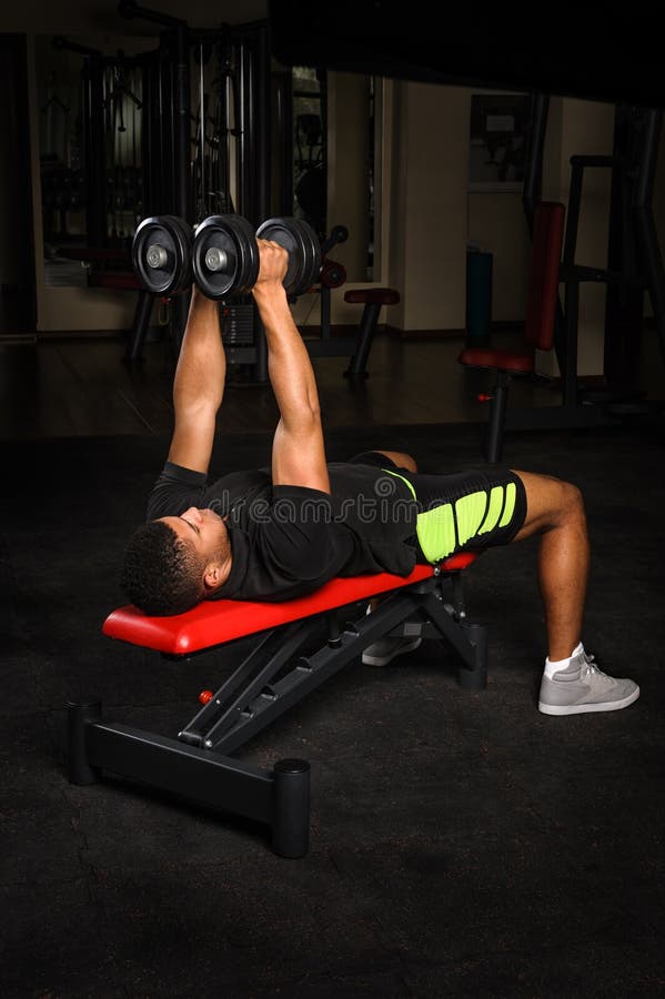 Le jeune homme faisant le banc de bras pilote la séance d'entraînement dans le gymnase photographie stock Le jeune homme faisant le banc de bras pilote la séance d'entraînement dans le gymnase photographie stock