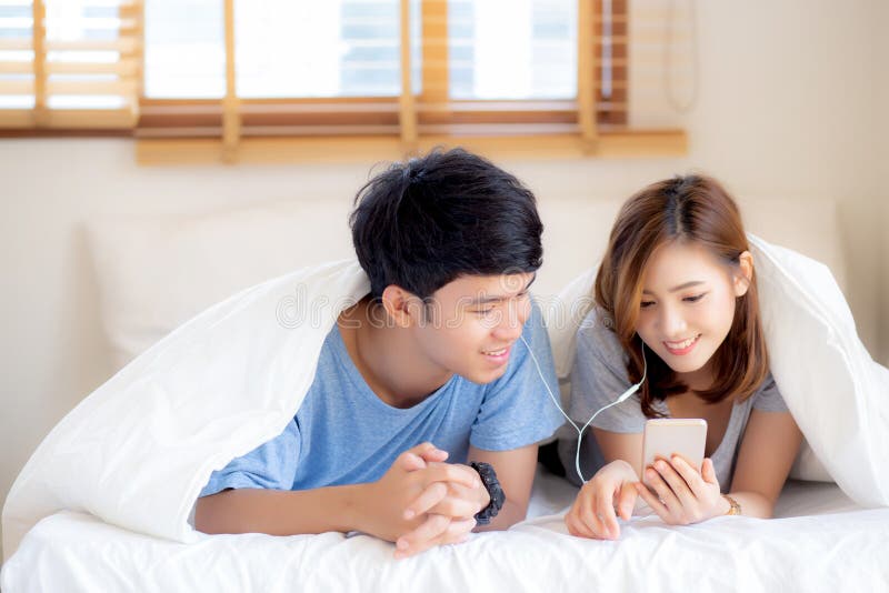 Le jeune sourire asiatique de couples écoutent musique avec le téléphone portable intelligent sur le lit à la chambre à coucher a photos stock Le jeune sourire asiatique de couples écoutent musique avec le téléphone portable intelligent sur le lit à la chambre à coucher a photos stock