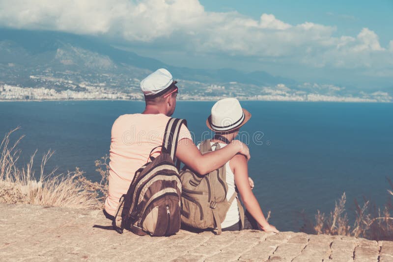 Le jeunes père et fils avec des sacs à dos s'asseyent sur la plage contre le contexte de la mer Concept de voyage de famille photos stock Le jeunes père et fils avec des sacs à dos s'asseyent sur la plage contre le contexte de la mer Concept de voyage de famille photos stock