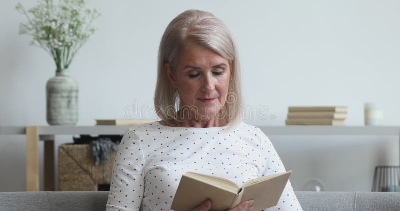 Le livre de lecture âgé moyen de femme assis sur le sofa banque de vidéos