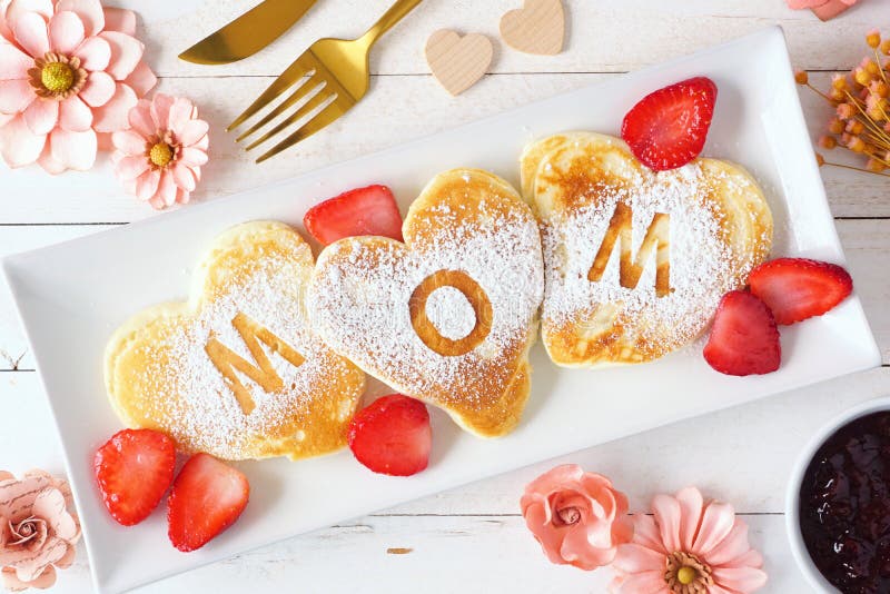 Le mamme diurne fanno i pancake a forma di cuore a colazione con le lettere della mamma sulla scena del tavolo sul legno bianco