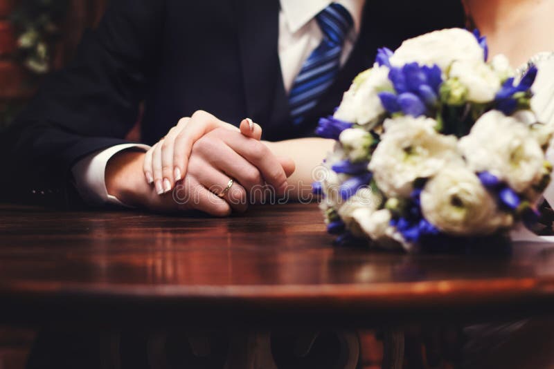Le mani delle persone appena sposate con le fedi nuziali ed il mazzo del ` s della sposa sulla tavola fotografia stock Le mani delle persone appena sposate con le fedi nuziali ed il mazzo del ` s della sposa sulla tavola fotografia stock
