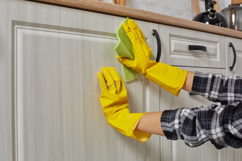 Le mani in guanti con lo straccio verde sta pulendo gli armadi da cucina - concetto del governo della casa e di lavoro domestico