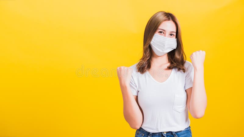 Asian beautiful happy young woman wearing face mask protection filter dust pm2.5, COVID virus and air pollution her raise hands glad excited cheerful after recovering from illness on yellow background. Asian beautiful happy young woman wearing face mask protection filter dust pm2.5, COVID virus and air pollution her raise hands glad excited cheerful after recovering from illness on yellow background