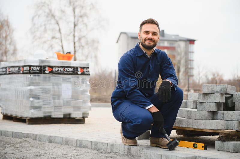 Le maître dans les gants pose des pavés en couches. Jetage de dalles de béton gris dans la cour de la maison sur le sable image stock