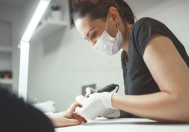 Professional manicure master dressed black in safety face mask using Electric Nail Polisher Tool for Glazing treatment manicure procedure. Everyday working people working concept. Professional manicure master dressed black in safety face mask using Electric Nail Polisher Tool for Glazing treatment manicure procedure. Everyday working people working concept