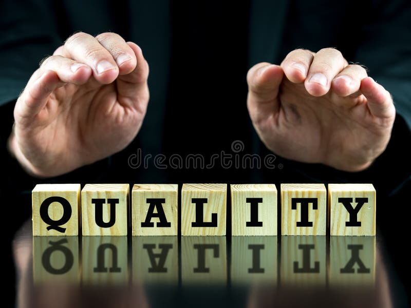 The word - Quality - on wooden cubes on a black reflective surface with the hands of a businessman in a suit held protectively above the blocks sheltering them. The word - Quality - on wooden cubes on a black reflective surface with the hands of a businessman in a suit held protectively above the blocks sheltering them.