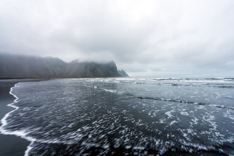 Le onde si schiantano a terra sulla spiaggia nera della penisola di Stokksnes con sullo sfondo la catena montuosa vestrahorn fotografia stock libera da diritti Le onde si schiantano a terra sulla spiaggia nera della penisola di Stokksnes con sullo sfondo la catena montuosa vestrahorn fotografia stock libera da diritti