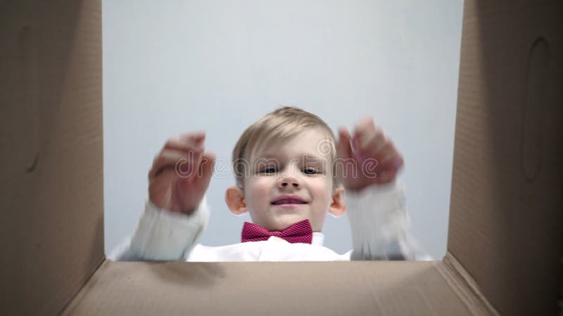 Le petit garçon blond heureux dans une chemise blanche avec un noeud papillon rouge regarde dans la boîte, est étonné et heureux