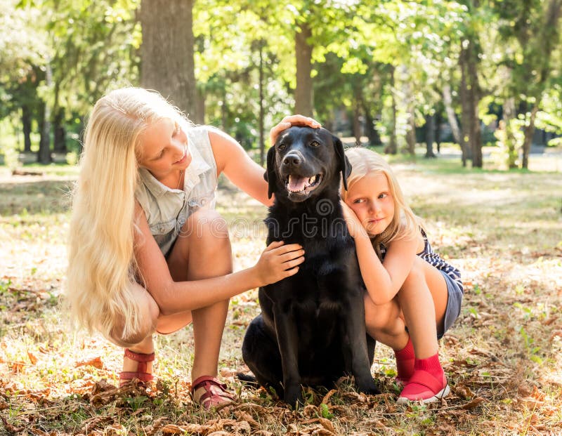 Le piccole ragazze bionde si siedono abbracciando un cane nero in un parco immagini stock Le piccole ragazze bionde si siedono abbracciando un cane nero in un parco immagini stock