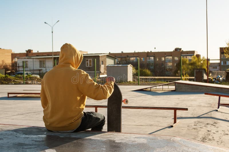 Le plan rapproché d'un adolescent s'est habillé dans un hoodie de jeans se reposant en parc de patin et tenant une planche à roul