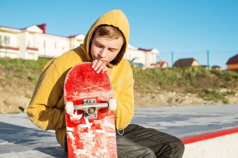 Le plan rapproché d'un adolescent s'est habillé dans un hoodie de jeans se reposant en parc de patin et tenant une planche à roul