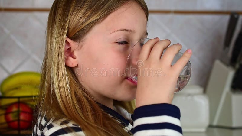 Le portrait de la petite fille blonde boit l'eau du verre dans le mouvement kitchenslow. banque de vidéos