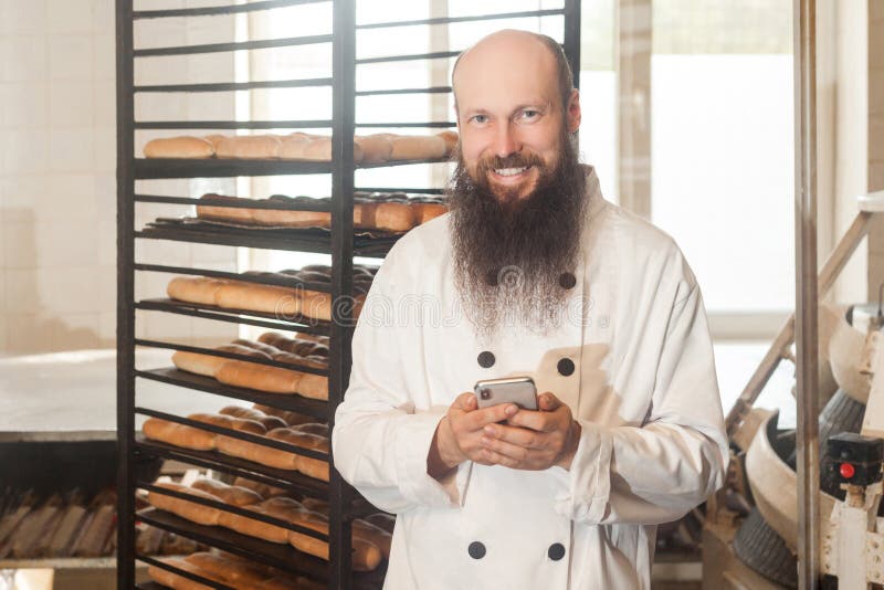 Portrait of satisfied young adult businessman baker with long beard in white uniform standing in bakery and have online order by phone, using phone with toothy smile Indoor, looking at camera. Portrait of satisfied young adult businessman baker with long beard in white uniform standing in bakery and have online order by phone, using phone with toothy smile Indoor, looking at camera