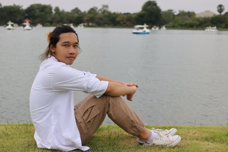 Le portrait du jeune homme asiatique s'asseyant sur une herbe verte en rivière de nature détendent le temps images stock Le portrait du jeune homme asiatique s'asseyant sur une herbe verte en rivière de nature détendent le temps images stock