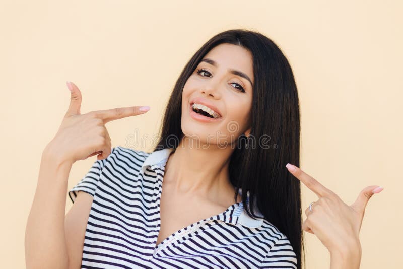 Le portrait du modèle femelle magnifique heureux indique avec les doigts antérieurs à son visage, fait de la publicité des accola photographie stock Le portrait du modèle femelle magnifique heureux indique avec les doigts antérieurs à son visage, fait de la publicité des accola photographie stock